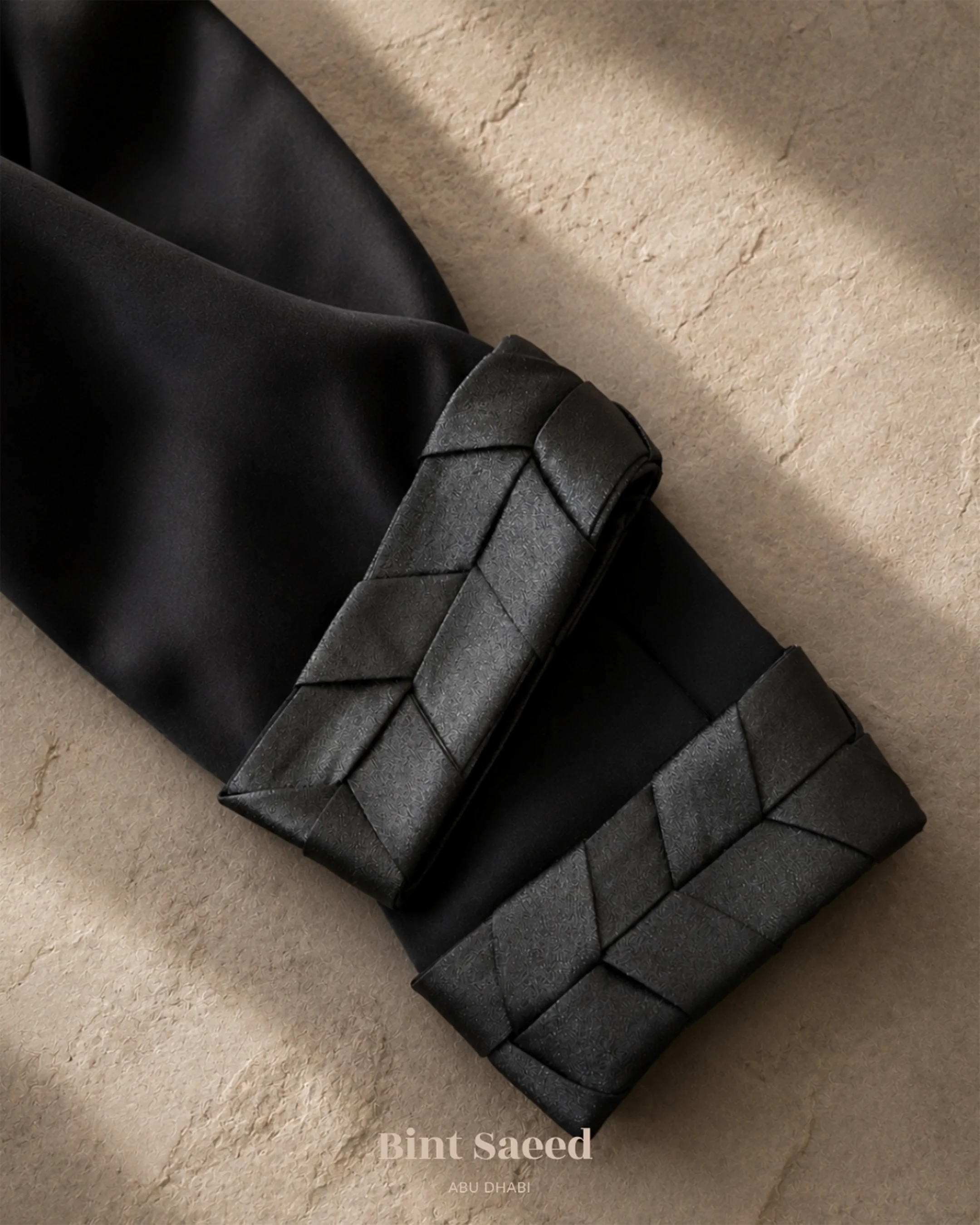 Detail manset Abaya Kensington dari Bint Saeed Abu Dhabi, Uni Emirat Arab, menampilkan trim tenun khas terinspirasi Al Khous, seni tradisional Emirati dalam menganyam pelepah palem, yang diinterpretasikan melalui anyaman organza glitter hitam yang halus. Dirancang di Abu Dhabi, Uni Emirat Arab. Pengiriman ke seluruh dunia.