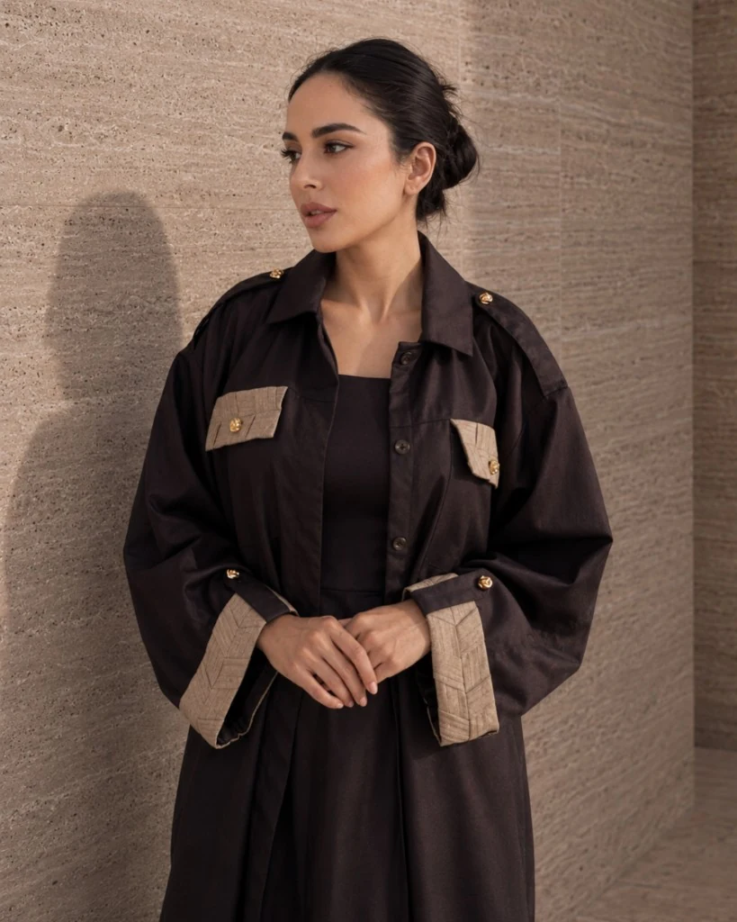 Potret gaya hidup Knightsbridge Khous Jacket Abaya Bint Saeed dalam Dark Brown — separuh badan di hadapan dinding batu, tangan bersilang, mempamerkan butiran tenunan terinspirasi Khous pada poket dada dan cuff dengan butang emas Knotted Line signature. Outerwear sopan Emirati kontemporari dan kraf warisan Al Khous dari Abu Dhabi, Emiriah Arab Bersatu. Dihasilkan di Abu Dhabi. Penghantaran ke seluruh dunia. Bint Saeed Abu Dhabi, Emiriah Arab Bersatu.