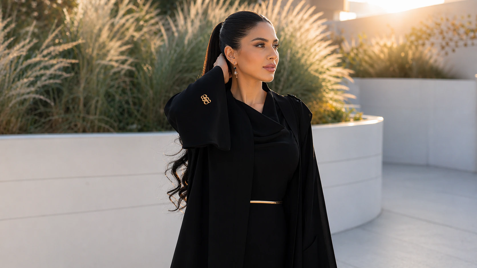 Bint Saeed Park Lane Abaya in Deep Black — luxury Emirati designer abaya with a draped shoulder scarf and wide cuffs fastened by the gold-tone Bint Saeed Monogram cufflink, worn at golden hour on a sunlit terrace. Bint Saeed Abu Dhabi, United Arab Emirates.