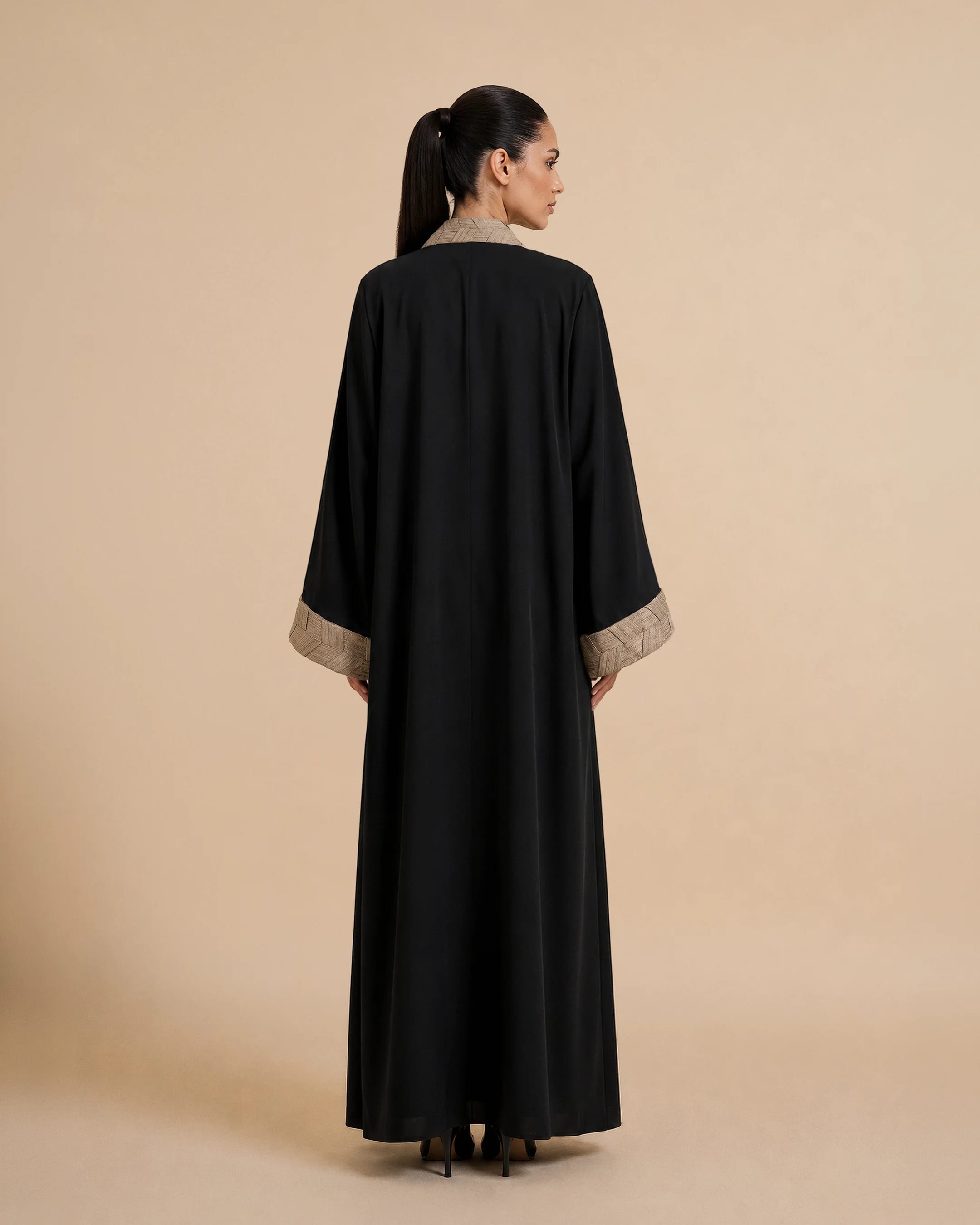 Abaya Belgravia dalam Hitam Pekat, tampak belakang. Abaya desainer dari Bint Saeed Abu Dhabi, Uni Emirat Arab, menonjolkan garis bersih, draperi panjang penuh, dan trim tenun tangan terinspirasi tradisi anyaman Khous Emirati.