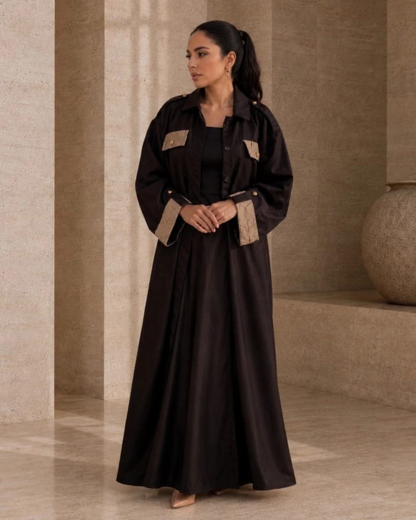 Knightsbridge Abaya Jacket Dark Brown — lifestyle | Khous | Bint Saeed Abu Dhabi Gambar lifestyle full-length Knightsbridge Khous Jacket Abaya Bint Saeed dalam Dark Brown — siluet jaket terbuka di interior batu, menampilkan detail tenun terinspirasi Khous pada saku dada dan manset dengan kancing emas Knotted Line signature. Outerwear modest Emirati kontemporer dan kerajinan warisan Al Khous dari Abu Dhabi, Uni Emirat Arab. Dibuat di Abu Dhabi. Pengiriman ke seluruh dunia. Bint Saeed Abu Dhabi, Uni Emirat Arab.