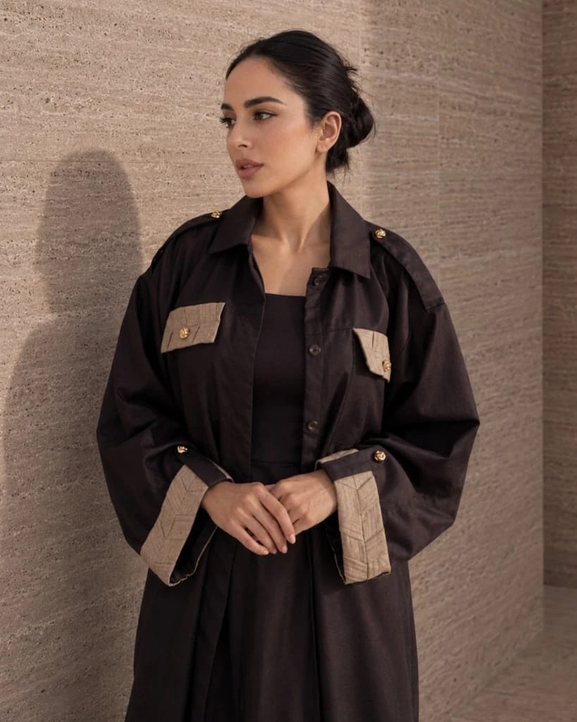 Knightsbridge Abaya Jacket Dark Brown — potret lifestyle | Khous | Bint Saeed Abu Dhabi Potret lifestyle Knightsbridge Khous Jacket Abaya Bint Saeed dalam Dark Brown — setengah badan di depan dinding batu, tangan tersilang, menampilkan detail tenun terinspirasi Khous pada saku dada dan manset dengan kancing emas Knotted Line signature. Outerwear modest Emirati kontemporer dan kerajinan warisan Al Khous dari Abu Dhabi, Uni Emirat Arab. Dibuat di Abu Dhabi. Pengiriman ke seluruh dunia. Bint Saeed Abu Dhabi, Uni Emirat Arab.