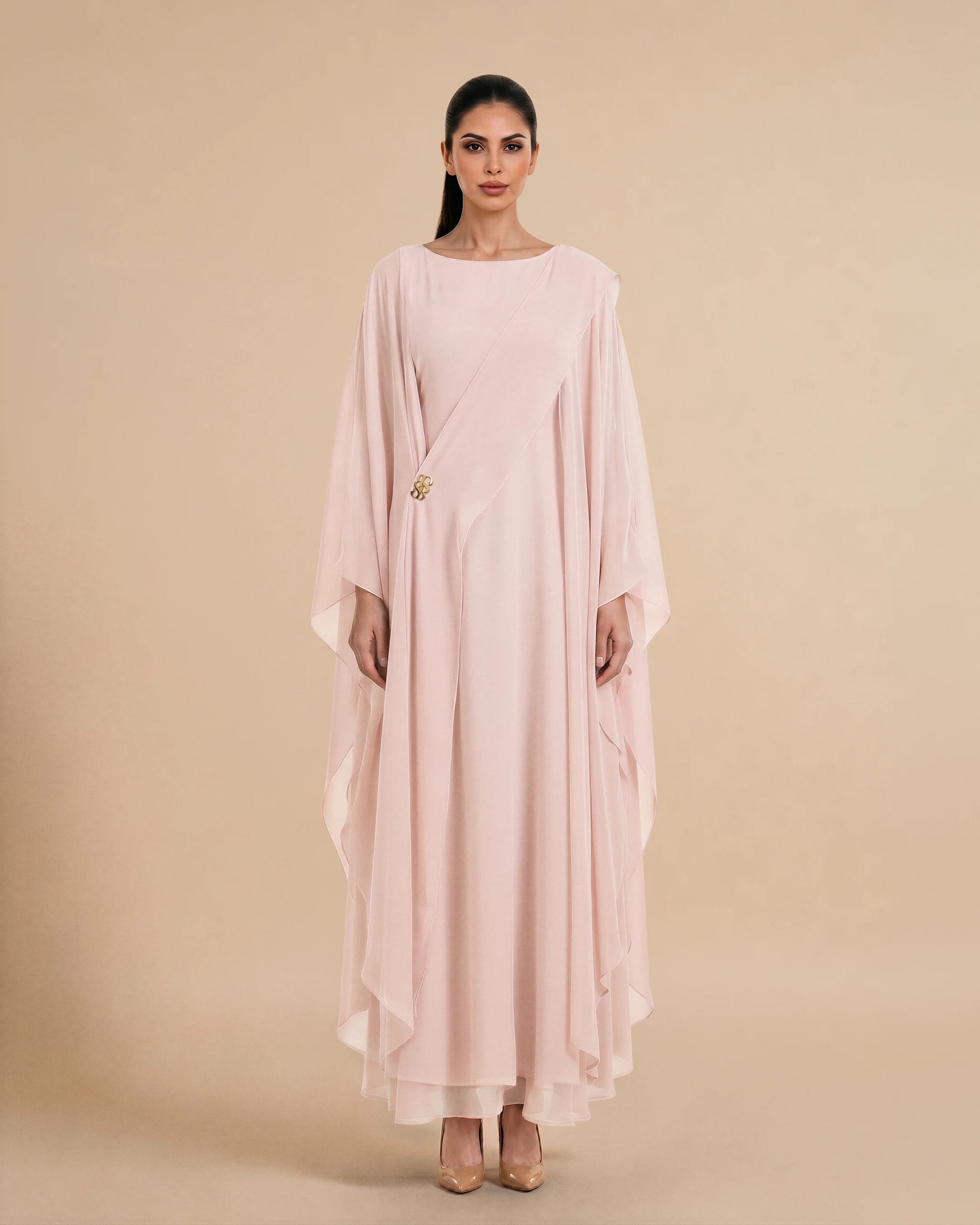 Kaftan Nothing Hill dalam kain chiffon Merah Muda Persik, tampak depan. Kaftan mewah wanita untuk acara dari Bint Saeed Abu Dhabi, Uni Emirat Arab, menampilkan garis leher bateau, siluet mengalir, dan lambang emas khas.
