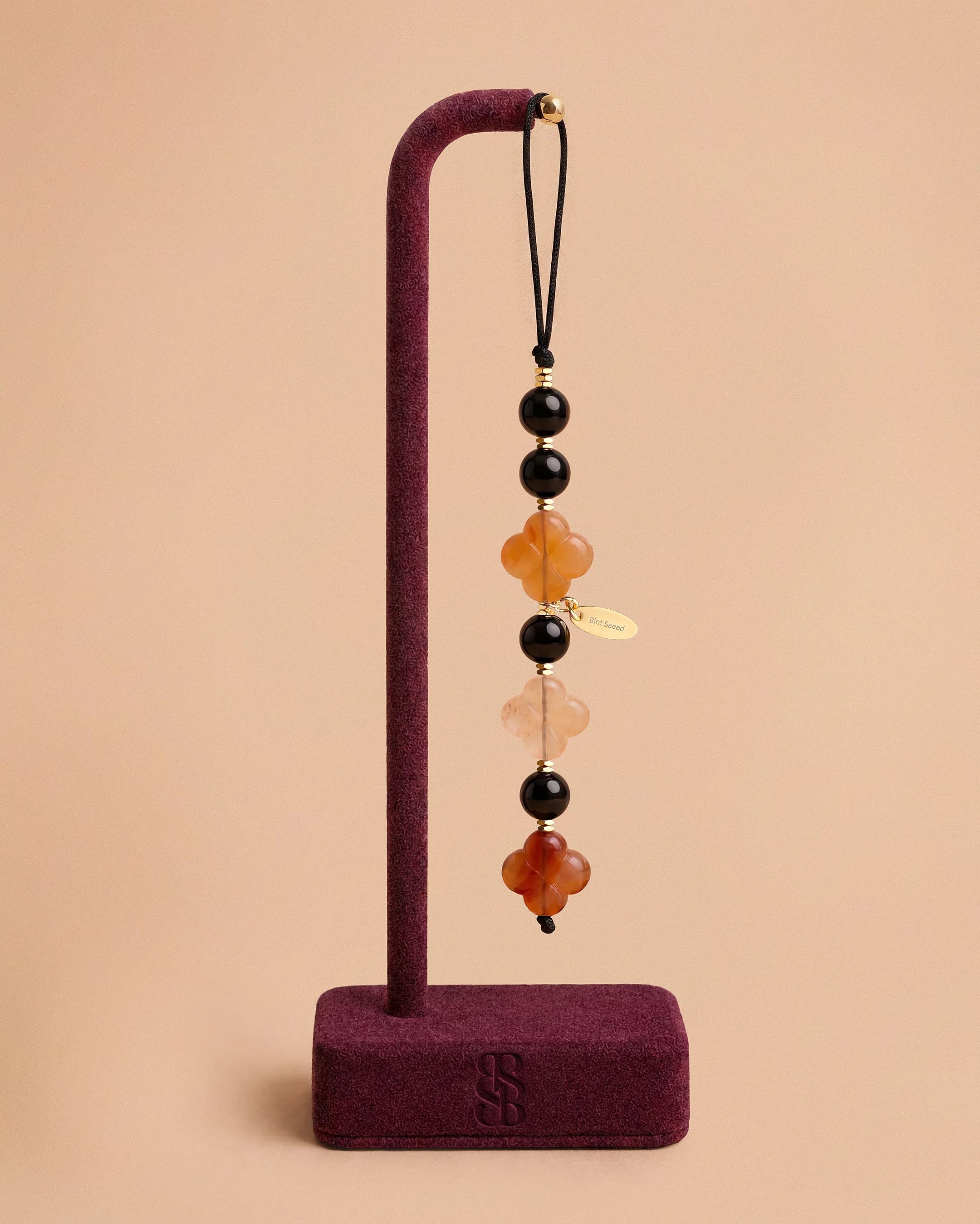Al Quaa Onyx natural stone phone charm by Bint Saeed: polished deep black onyx beads, hand-carved Carnelian Al Ain Rosette and gold-plated faceted hematite accents on display. Elegant luxury phone jewellery for lovers of natural stones and contemporary Emirati accessories. Made in Abu Dhabi, United Arab Emirates. Worldwide shipping. Bint Saeed Abu Dhabi, United Arab Emirates.