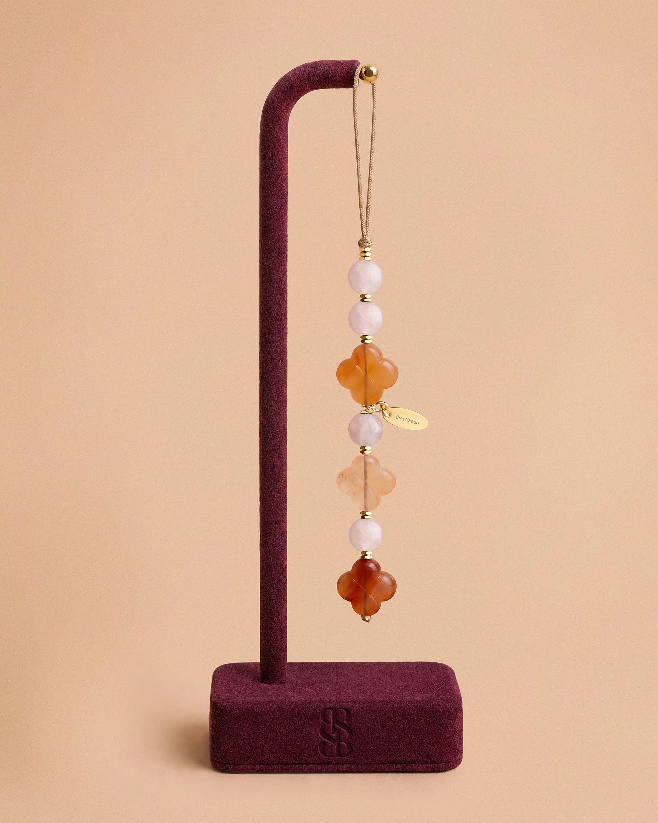 Al Quaa Rose Quartz natural stone phone charm by Bint Saeed: soft blush rose quartz beads, hand-carved Carnelian Al Ain Rosette and gold-plated faceted hematite accents on display. Romantic luxury phone jewellery for lovers of natural stones, phone charms and refined accessories. Made in Abu Dhabi, United Arab Emirates. Worldwide shipping. Bint Saeed Abu Dhabi, United Arab Emirates.