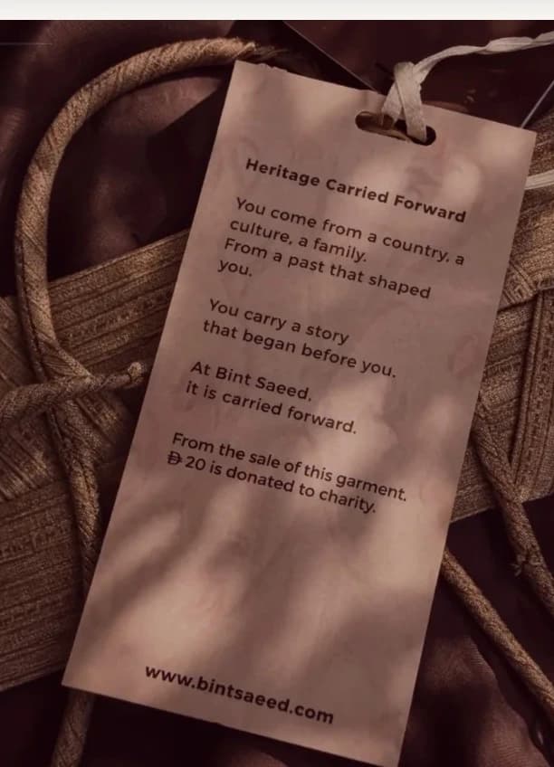 Giving Forward hangtag — Heritage Carried Forward charity swing tag on luxury fabric, with 20 AED from each garment donated to charity. Bint Saeed Abu Dhabi, United Arab Emirates.