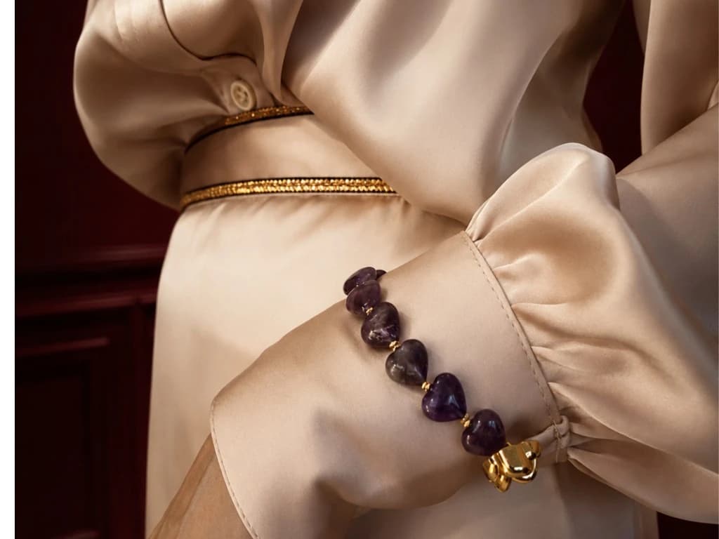 Bint Saeed Signature Strand with heart-shaped amethyst stones and gold accents, attached to the cuff of a champagne silk blouse. Bint Saeed Abu Dhabi, United Arab Emirates.